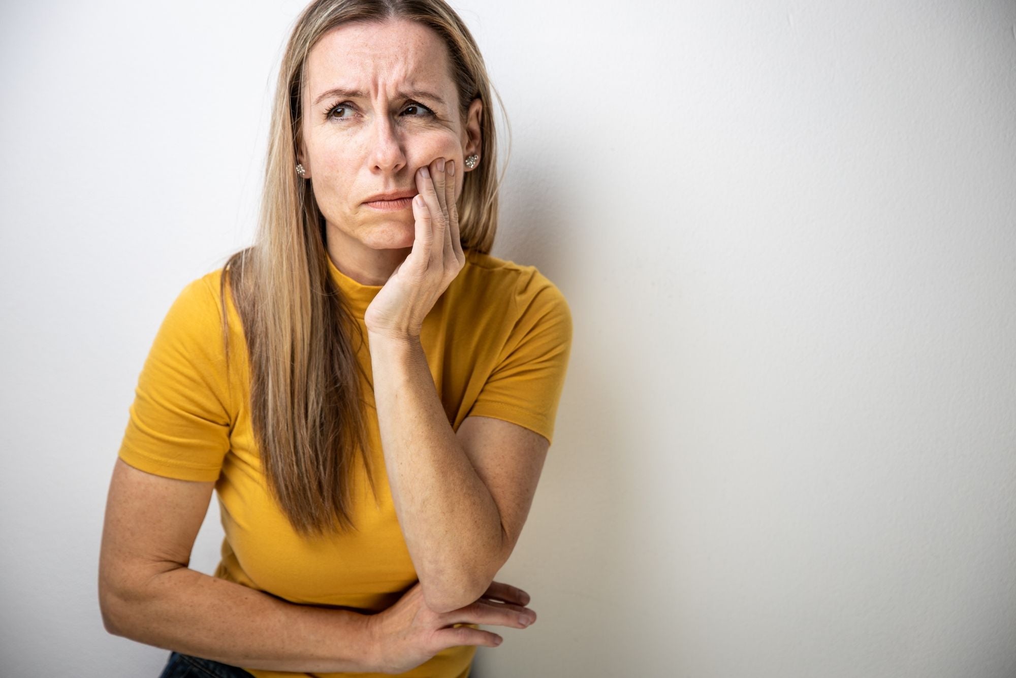 A blonde woman wearing a yellow shirt holds her face in pain against a white background.