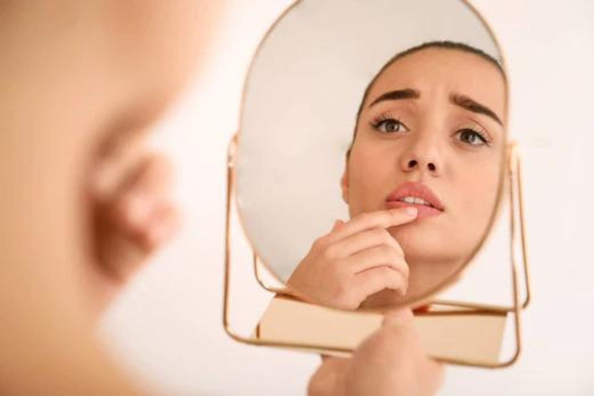 A woman worried about the herpes stigma looks in a handheld mirror while touching her face.