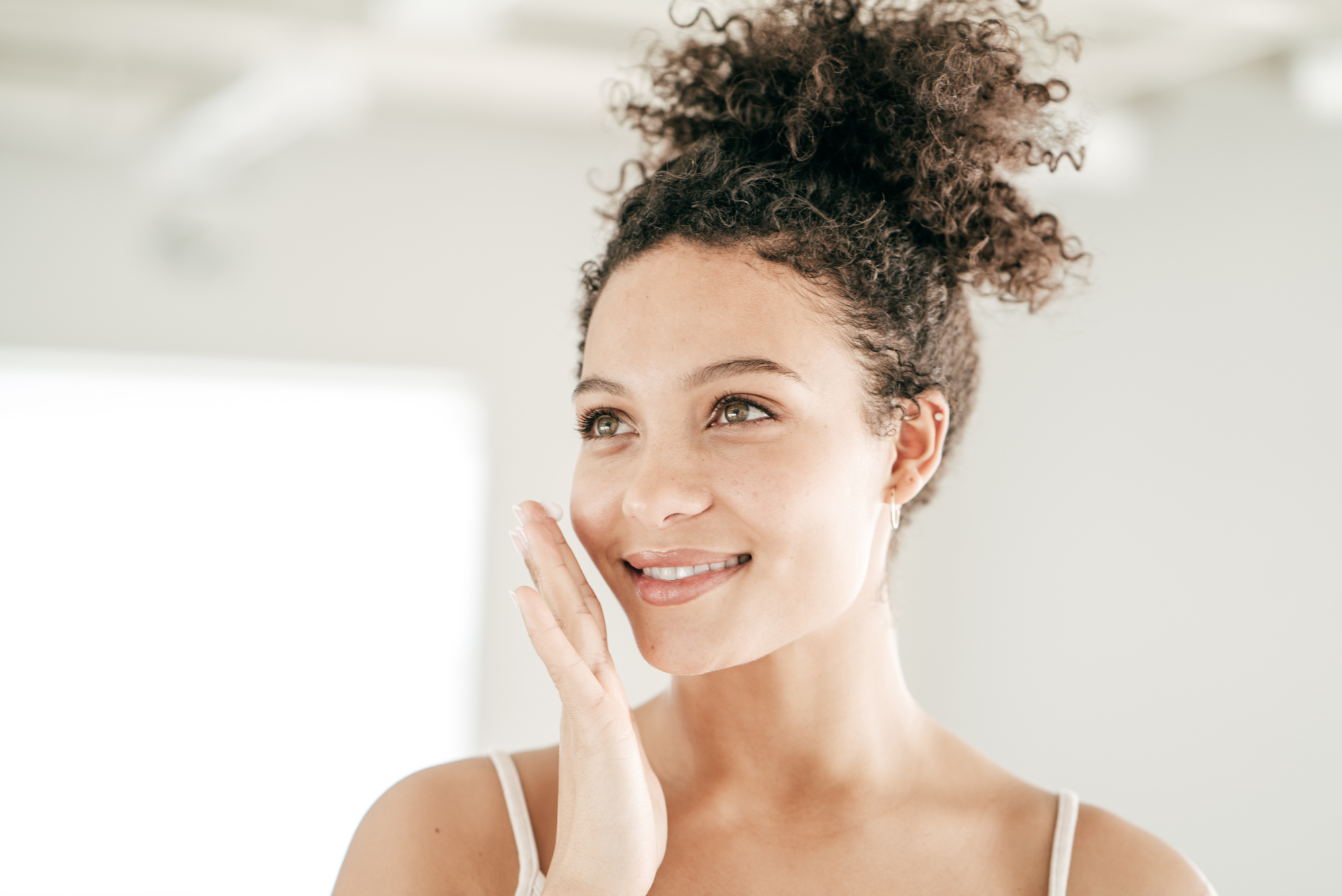 A woman in a white tank top smiles as she applies retinol to her face after using red light therapy at home.