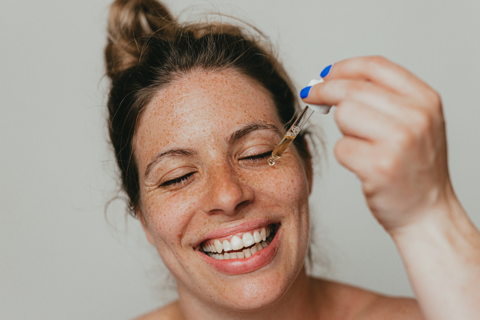 A woman with many freckles and blue fingernails smiles widely as she applies serum to her cheek before red light therapy.